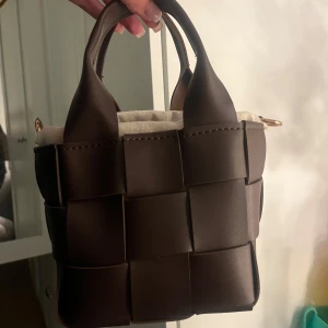 Brun flätad handväska med axelrem - Säljer en brun handväska i flätad design med både handtag och justerbar axelrem i skinnimitation. Väskan har en innerpåse i beige bomull med snörning och detaljer i guld. Perfekt storlek för det viktigaste och riktigt trendig look.