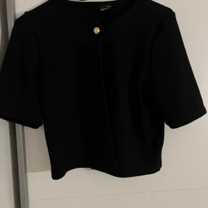 Svart kofta från Gina Tricot XS - Stilren svart kofta från Gina Tricot i kortare modell med halvkort ärm och rund halsringning. Kavajen har en snygg guldig knapp framtill som detalj. 