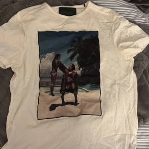 Vit t-shirt med strandmotiv - Vit t-shirt med ett stort tryck på framsidan som visar två personer vid en pool och palmer i bakgrunden. T-shirten har rund hals och korta ärmar. Perfekt för dig som gillar unika och konstnärliga prints.