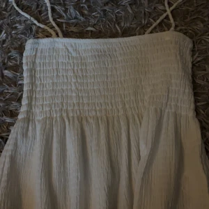 Vit T-shirt från Gina Tricot - Vit T-shirt från Gina Tricot i storlek M. T-shirten har smock upptill, tunna axelband och ett lätt, luftigt tyg som ger en skön känsla. Perfekt för varma dagar och har en enkel, stilren look som är lätt att matcha med accessoarer.