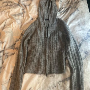 Grå ribbad zip hoodie med dragkedja - Mysig grå zip hoodie i ribbstickad ullmix. Tröjan har huva, långa ärmar och hel dragkedja framtill. Perfekt för lager-på-lager och chill dagar. Enkel och stilren design som funkar till det mesta. Tröjan har hål för tummarna och på ena armen har den rivits upp lite och blivit lite större men ingenting man lägger märke till så mycket. Pris kan diskuteras.