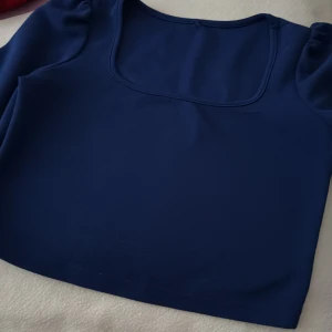 Marinblå croppad topp med puffärm - Snygg marinblå croppad topp med fyrkantig halsringning och puffade axlar. Perfekt för dig som gillar stilrena plagg med en twist. Materialet känns mjukt och stretchigt, vilket gör den bekväm att bära hela dagen.