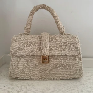 Beige handväska i ull med guldspänne - Snygg beige handväska med fluffig, stickad ullstruktur och ett elegant guldspänne framtill. 