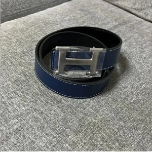 Mörkblå skärp i skinn - Snyggt mörkblått  bälte i skinn med en stor, silverfärgad H-spänne. Bältet har vita sömmar längs kanterna som ger en clean kontrast. Perfekt accessoar för att lyfta din outfit med en lyxig touch.