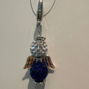 Halsband med blå och silver ängel - Unikt halsband med en ängelberlock i silverfärgad metall. Berlocken har en glittrig vit kula som huvud och en mörkblå glittrig kula som kropp, samt detaljerade vingar i metall. Perfekt för dig som gillar personliga smycken med lite extra bling.