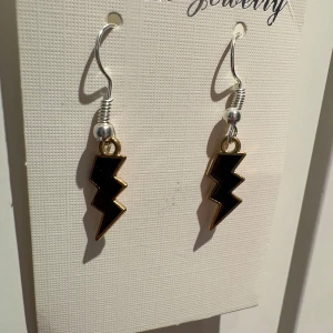 Svarta blixtformade örhängen - Coola örhängen i form av blixtar med svart insida och guldfärgad kant. Hängande modell med silvriga krokar som ger en edgy vibe. Perfekta för dig som vill sticka ut och addera lite attitude till din look.
