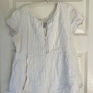 Vit blus från Odd Molly med broderi - Supersöt vit blus från Odd Molly med diskreta ränder, korta ärmar och v-ringning med virkade detaljer. På ryggen finns ett broderat Odd Molly-märke med fjädrar. Blusen är luftig och har en avslappnad passform, perfekt till jeans eller kjol.