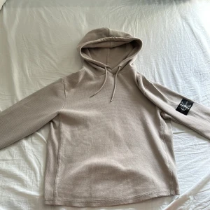 Beige hoodie från Calvin Klein Jeans - Säljer en beige hoodie från Calvin Klein Jeans med svart logotyp-patch på ärmen. Tröjan har lång ärm, huva med dragsko och är tillverkad i ett strukturerat bomullsmaterial. Perfekt för en avslappnad och stilren look.