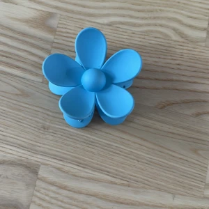 Blå blomformad hårklämma - Säljer en söt blå hårklämma i form av en blomma med sex rundade kronblad. Perfekt för att ge din frisyr en färgglad och lekfull touch. Klämman är gjord av plast och har en mjuk, matt finish.