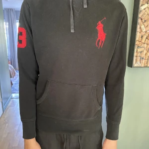 Svart hoodie från Polo Ralph Lauren - Svart hoodie från Polo Ralph Lauren med stor röd logga på bröstet och siffran 3 broderad på ärmen. Klassisk huva och magficka framtill. Mjuk insida och ribbade muddar vid ärmslut och nederkant. Perfekt för chill dagar eller när du vill ha en clean hoddie.
