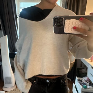 Beige croppad stickad tröja - Säljer en beige croppad stickad tröja med loose passform och lång ärm. Tröjan har en bred v-ringad hals som ger en chill offshoulder-look. Perfekt att matcha med svarta jeans för en avslappnad vibe.