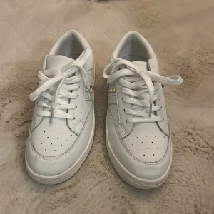 Vita sneakers från Sofie Schnoor   (Bara använda en gång) - Snygga vita sneakers från Sofie Schnoor med klassisk design, perforerad tå och coola guldfärgade nitar på sidan. Skorna har snörning och är gjorda i skinnliknande material. Perfekta för dig som gillar stilrena och trendiga detaljer.