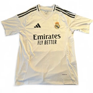 Real Madrid vit matchtröja Adidas M - Snygg Real Madrid fotbollströja från Adidas i slim fit, storlek M. Tröjan är vit med svarta detaljer och har klubbmärket och sponsortryck på bröstet. Tillverkad i lätt och ventilerande polyester med klassiska svarta ränder på axlarna. Perfekt för dig som älskar fotboll och vill ha en autentisk look.