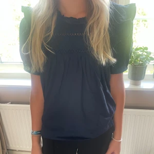 Mörkblå blus med volangärmar - Supersöt mörkblå blus med volangärmar och broderade detaljer upptill. Blusen har en lös passform och korta ärmar med extra volym. Perfekt för dig som gillar romantisk och trendig stil.