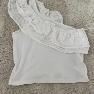 Vit oneshoulder topp med volang Gina Tricot - Supersöt vit oneshoulder topp från Gina Tricot med bred broderad volang och tunna axelband. Toppen är i mjuk bomull och har ett romantiskt hålmönster på volangen. Perfekt för sommarens alla tillfällen och ger en fräsch vibe.