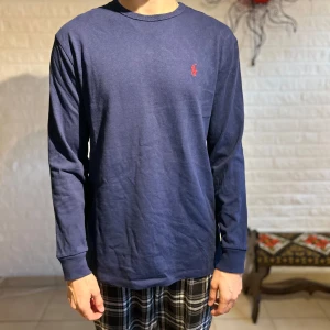 Mörkblå långärmad tröja Polo Ralph Lauren - Snygg mörkblå långärmad tröja från Polo Ralph Lauren i klassisk passform. Tröjan har en rund hals och är tillverkad i mjuk bomull. På bröstet finns den ikoniska röda Polo-loggan broderad. Perfekt för dig som gillar stilrena och tidlösa plagg. Endast använd en gång, 