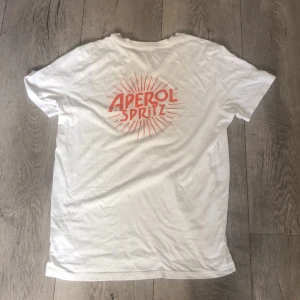 Vit Aperol Spritz t-shirt - Vit t-shirt med korta ärmar och rund hals. På framsidan finns ett rött 'A' tryck och på ryggen ett stort Aperol Spritz-tryck i rött med solstråle-design. T-shirten är gjord i mjuk bomull och har en clean, chill vibe som passar perfekt till sommaren. Inte jag som har skrivit texten där uppe Hahah men tröjan är det iallafall o storlek S och M men tröjan är fet som fan men lite smutsig men den går o tvätta såklart. Jag tog storlek 42 men den är i S och M