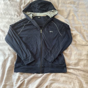 Hugo Boss zip  - Svart zip hoodie från Hugo Boss med dragkedja framtill och broderad logga på bröstet. Zip hoodien har huva med ljusgrå insida, långa ärmar och gråa detaljer längs axlarna. Tillverkad i mjuk bomull, perfekt för chill dagar. Pris kan diskuteras vid snabb affär 