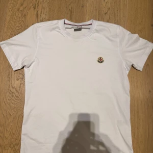 Vit Moncler t-shirt med logga - Vit t-shirt från Moncler i mjuk bomull med klassisk rund hals och korta ärmar. På bröstet sitter Monclers ikoniska broderade logga i rött, blått och gult. Enkel och clean design som funkar till jeans eller shorts. Storlek S men sitter som M. Nytt skick och knappast använd.