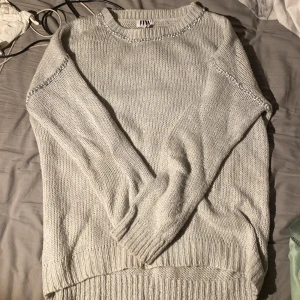 Ljusgrå stickad tröja med pärldetaljer - Säljer en ljusgrå stickad tröja från JW med kedjedetaljer vid halsringning och axlar. Tröjan har rund hals, långa ärmar och breda ribbade muddar vid ärmslut och nederkant. Perfekt för dig som gillar stilrena och mysiga plagg.