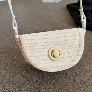 Beige axelväska i bomull med twistlås - Söt beige axelväska i vävd bomull med rundad form och twistlås i plast framtill. Väskan har ett vitt innerfoder med dragkedjefack och smal axelrem. Perfekt storlek för mobil och småprylar, enkel att matcha till olika outfits.