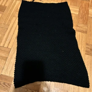 Svart stickad halsduk  - Svart stickad kjol med enkel, rak form. Materialet är grovstickat och ger en mysig känsla. Kjolen är enfärgad och har en klassisk look som funkar till många olika stilar. Perfekt för dig som gillar handgjorda plagg och vill ha något unikt i garderoben.