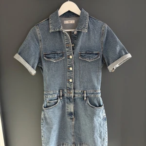 Kort jeansklänning från mango Denim - Snygg kort jeansklänning från mango Denim med klassisk skjortkrage, korta ärmar och knappar framtill. Klänningen har fickor både fram och bak samt bälteshällor i midjan. Perfekt för en cool och avslappnad look.