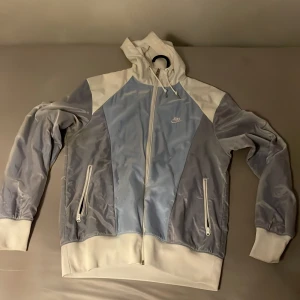 Nike vindjacka i blått och vitt - Nike vindjacka med huva i ljusblått och vitt, stora loggan på ryggen och mindre logga fram. Jackan har dragkedja, två fickor med dragkedja och ribbade muddar. Perfekt för dig som gillar sportig stil och vill sticka ut med snygga färgblock.