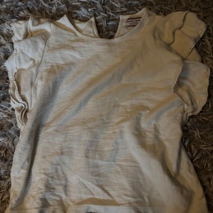 Vit volangblus från Holly & Whyte - Supersöt vit blus från Holly & Whyte med volangdetaljer på ärmarna. Blusen har rund halsringning och är tillverkad i ett mjukt, lätt material som känns skönt mot huden. Perfekt för dig som gillar en feminin och stilren look.