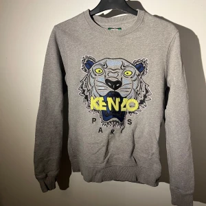 Grå sweatshirt med tiger från Kenzo - Säljer en grå sweatshirt från Kenzo med ikonisk broderad tiger på bröstet och färgglada detaljer i blått, gult och svart. Tröjan har rund hals, ribbade muddar och lång ärm. Perfekt för dig som gillar streetstyle och vill sticka ut.