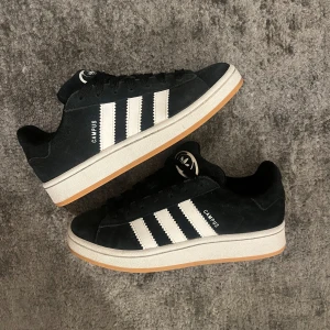Svarta adidas campus - Säljer ett par klassiska Adidas Campus sneakers i svart mocka med vita ränder och gummisula. Skorna har både svarta och vita skosnören så du kan variera stilen. Ikonisk Adidas-logga på plösen och Campus-tryck på sidan. Perfekta för en clean och tidlös look. Skickar med de vita snörena vid sidan av så kan man byta som man vill 💕