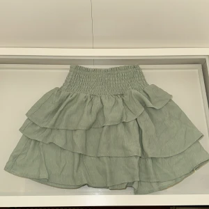 Grön volangkjol från H&M - Superfin ljusgrön kortkjol från H&M med bred smock i midjan och flera lager volanger. Kjolen är i ett mjukt, tunt tyg som känns luftigt och passar perfekt till varmare dagar. Enkel att matcha med både t-shirt och hoodie.