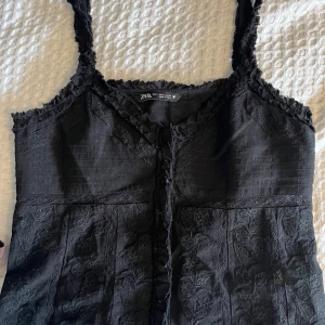 Svart blommig linnetopp från Zara - Supersnygg svart linnetopp från Zara med blommigt broderi och spetsdetaljer. Toppen har v-ringning, smala axelband och fransad kant längs halsen. Perfekt att styla med jeans eller kjol för en cool look.