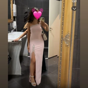 Ljusrosa lång klänning med slits strl S - Säljer en elegant ljusrosa långklänning med en glittriga detaljer längs halsringningen och slitsen. Klänningen har en hög slits framtill och figurnära passform som framhäver siluetten. Perfekt för bal eller fest!