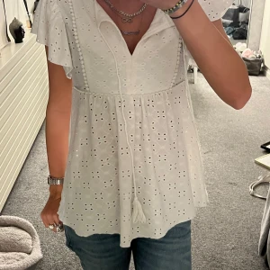 Vit blus  - Superfin vit blus från SHEIN i storlek XS med broderade hålmönster, v-ringning och volangärmar. Blusen har knytband med tofs framtill och en luftig, flowy passform. Perfekt för varma dagar och ger en romantisk vibe.