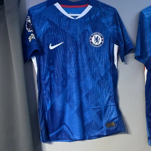 Chelsea hemmatröja Nike Delap #9 - Chelsea FC hemmatröja i blått med Delap och nummer 9 på ryggen. Tröjan har korta ärmar, vit V-ringning och diskret geometriskt mönster. Premier League-märke och 'No room for racism' på ärmen. Tillverkad i lätt och ventilerande material.