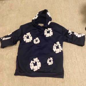 Blå hoodie från Denim Tears med tryck - Säljer en mörkblå hoodie från Denim Tears med unika vita tryck över hela tröjan. Tröjan har huva och känguruficka framtill. Perfekt för dig som vill sticka ut med en streetwear-look. Materialet känns mjukt och skönt mot huden.