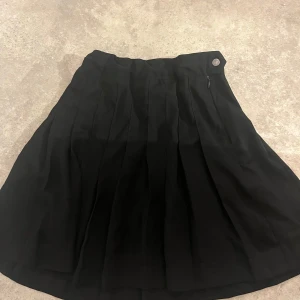 Svart plisserad kjol från Lindex - Snygg svart plisserad kjol från Lindex med dragkedja och knapp i midjan. Kjolen har en klassisk A-linjeform och är tillverkad i ett mjukt polyestertyg som ger ett stilrent intryck. Perfekt för dig som gillar enkel och tidlös stil.