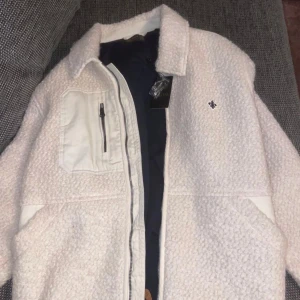 Vit teddyjacka från Morris Stockholm - Säljer en snygg vit teddyjacka från Morris Stockholm, modell Brookley Pile Jacket. Jackan har en oversized look, dragkedja framtill, stora fickor och detaljer i vitt tyg på bröstficka och axelparti. Perfekt för kalla dagar och riktigt trendig just nu.