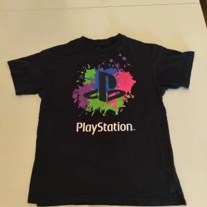 Svart PlayStation t-shirt med färgstänk - Svart t-shirt från PlayStation med stort tryck på bröstet. Trycket har färgstänk i rosa, lila, blått och grönt runt den klassiska PlayStation-loggan. T-shirten har rund hals och korta ärmar, tillverkad i mjuk bomull. Perfekt för dig som älskar gaming och vill sticka ut. Storlek: 158/164 cm (12-14 år)