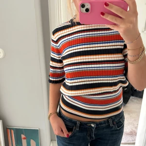 Randig ribbad topp i retrostil - Säljer en färgglad ribbad topp med randigt mönster i svart, orange, vit, blå och röd. Modellen har korta ärmar och sitter tajt där nere!