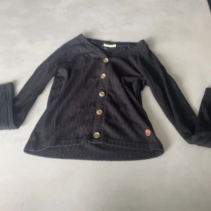 Svart ribbad blus med knappar - Svart ribbad blus från Ü i storlek 146/152. Toppen har lång ärm, v-ringning och snygga träfärgade knappar framtill. Materialet är mjukt och stretchigt, perfekt för dig som gillar enkel men stilren look. Liten rund logodetalj nertill. Knapparna går inte att knäppa upp!
