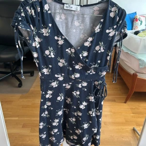 Blommig kortklänning från Hollister - Supersöt kortklänning från Hollister i marinblått med ljusa blommor. Klänningen har omlott-v-ringning, knytband i midjan och korta ärmar med snörning. Perfekt för sommarens alla tillfällen, mjukt bomullsmaterial och skön passform.