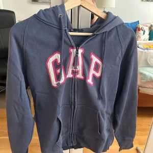 Mörkblå GAP hoodie med dragkedja - Mörkblå hoodie från GAP i storlek M med vit och rosa broderad logga framtill. Tröjan har huva med snörning, dragkedja och två fickor. Perfekt för chill dagar och enkel att matcha med jeans eller mjukisbyxor.