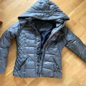 Grå quiltad dunjacka Rockandblue - Snygg grå dunjacka från Rockandblue med quiltat mönster, huva och flera dragkedjefickor. Jackan har Rockandblue-märke på ärmen och är perfekt för kalla dagar. Stängs med dragkedja och tryckknappar. Passar dig som gillar stilren och sportig look.