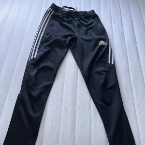 Svarta Adidas träningsbyxor  - Svarta träningsbyxor från Adidas med klassiska vita ränder längs sidorna och logga på låret. Byxorna har snörning i midjan och dragkedjor vid bensluten. Perfekta för sport eller chill. Vid frågor hör av dig!