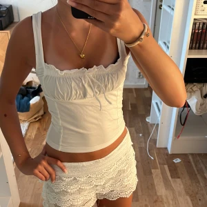 Vit linnetopp med rosett och rynk - Supersöt vit linnetopp med breda axelband, rynkade detaljer över bysten och en liten rosett framtill. Toppen är figurnära och har en croppad passform som passar perfekt till sommaren. Materialet känns mjukt och stretchigt, vilket gör den bekväm att bära hela dagen.