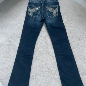 Mörkblå bootcut jeans från Gina Tricot - Snygga mörkblå jeans från Gina Tricot med bootcut passform och kontrastsömmar. Bakfickorna har broderade detaljer med bling och coolt mönster. Jeansen har justerbar midja på insidan och klassisk femficksdesign.