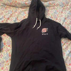 Svart hoodie med ros från Dope - Svart hoodie från Dope. Hoodien är använd och tvättad, därför syns lite små defekter på rosen på ryggen. 