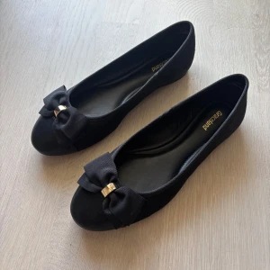 Svarta ballerinaskor med rosett från Graceland - Snygga svarta ballerinaskor från Graceland med rund tå och dekorativ rosett framtill, detaljerad med ett guldfärgat spänne. Skorna har en klassisk och stilren look, perfekt för dig som gillar enkel men söt stil. Materialet är mjukt och bekvämt.